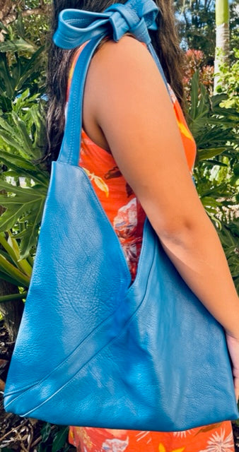 A woman holding on her shoulder a cobalt blue leather hobo bag with a tied strap against a tropical background. made in hawaii.