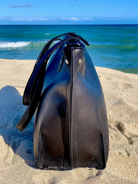 Onyx Black tote bag side view, on sand with ocean in the background.