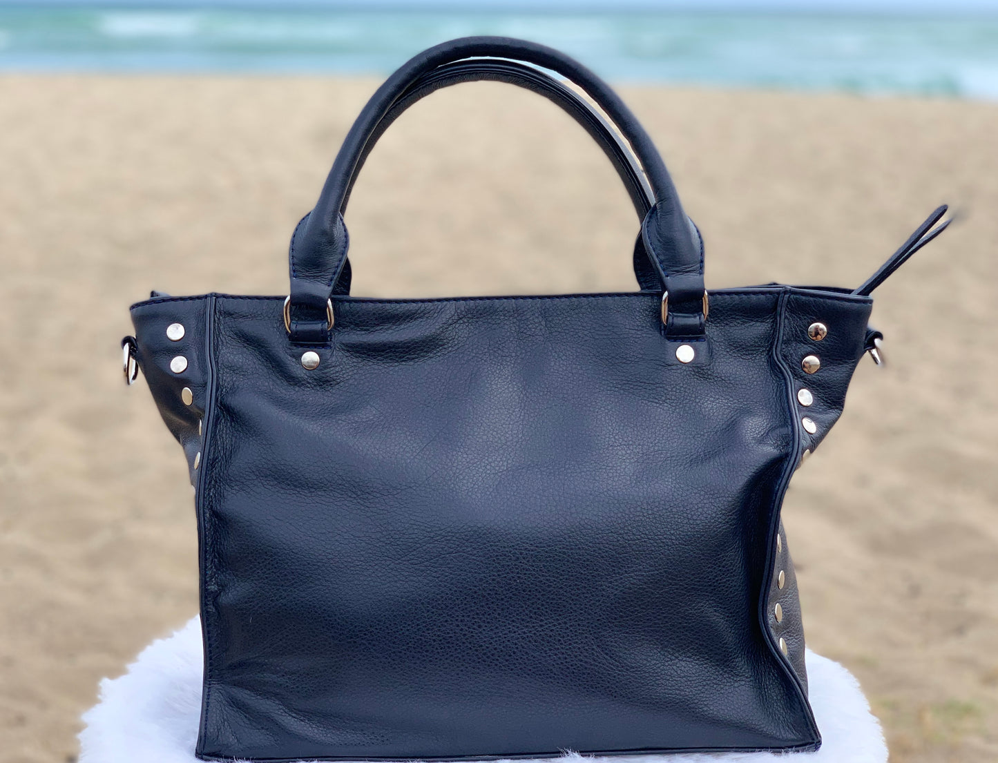 Sapphire leather satchel bag back view on a sandy beach with ocean in the background