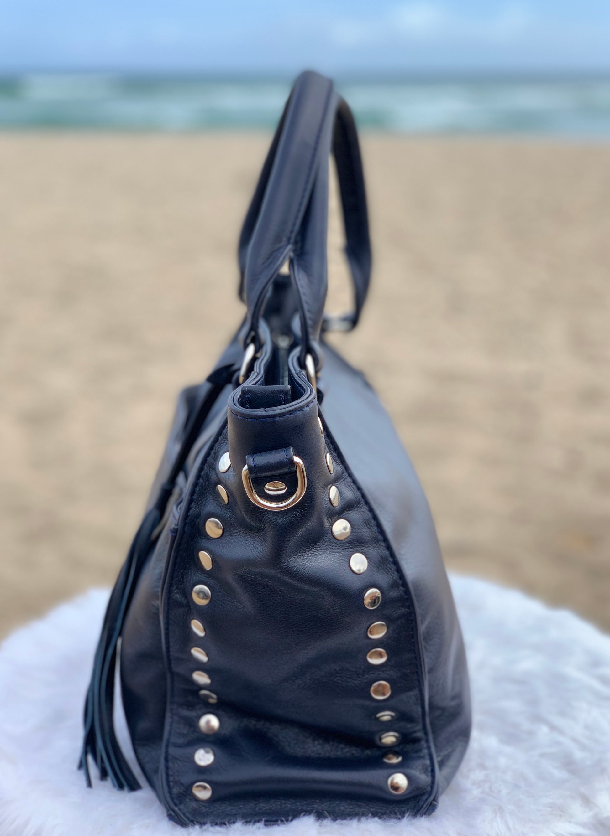 Sapphire leather satchel bag side view with gold studs and tassels on a fluffy white surface with a beach background