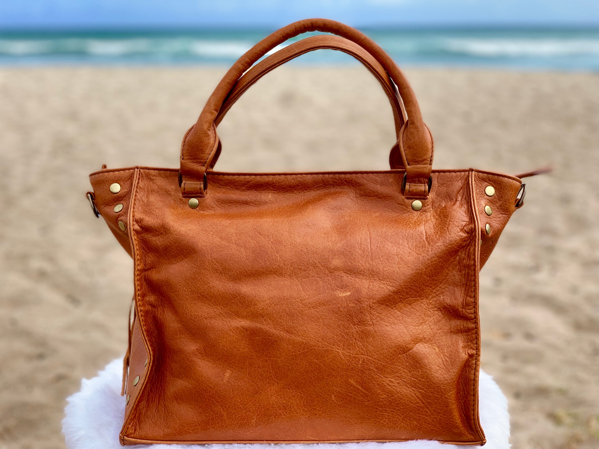 Whisky Brown leather satchel bag back view on a sandy beach with ocean in the background