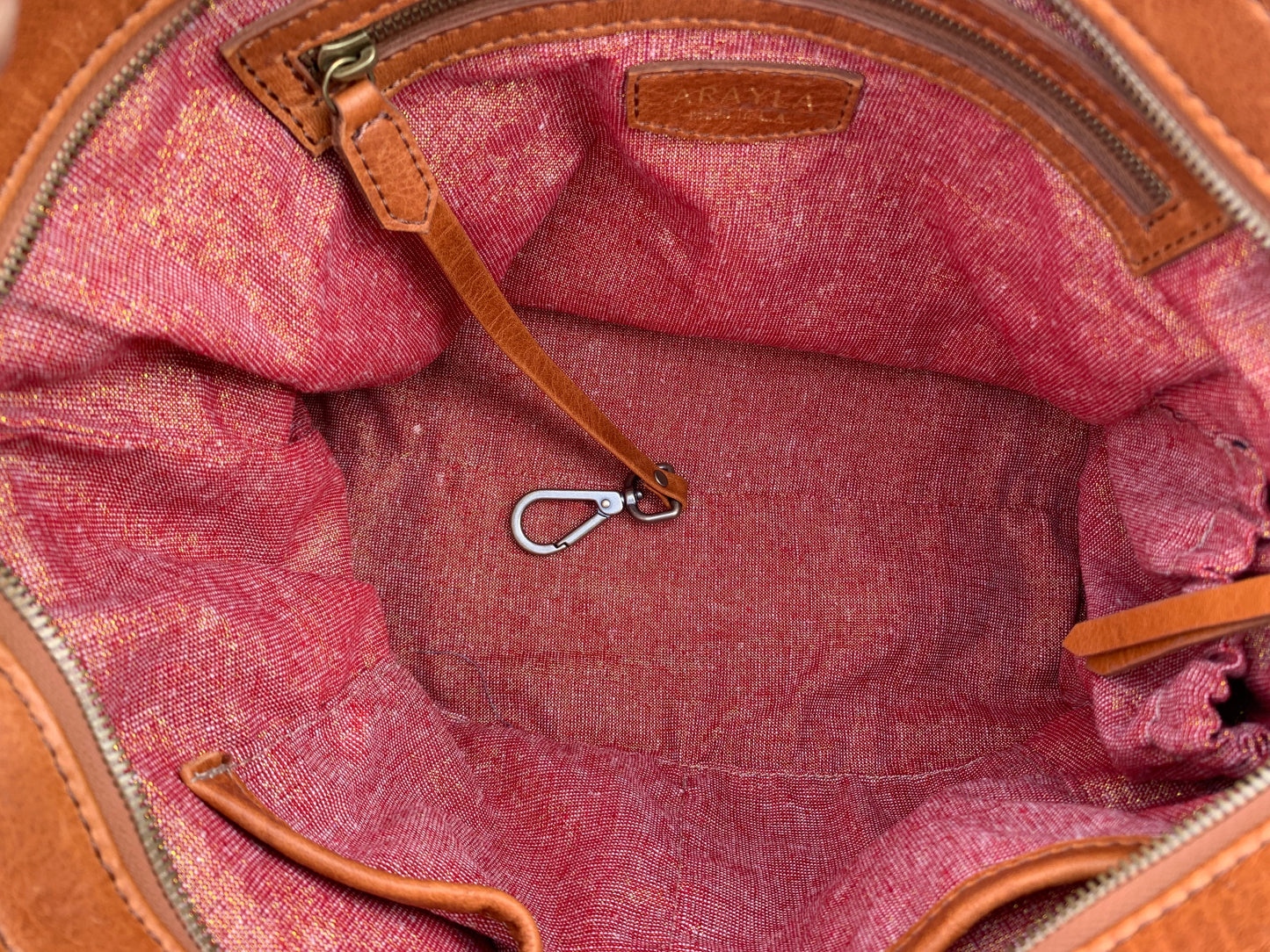 Close-up of the whisky brown satchel bag interior with red and golden shimmer textile lining, key leash, zip pocket, two slip mobile pockets and sunglasses pocket.