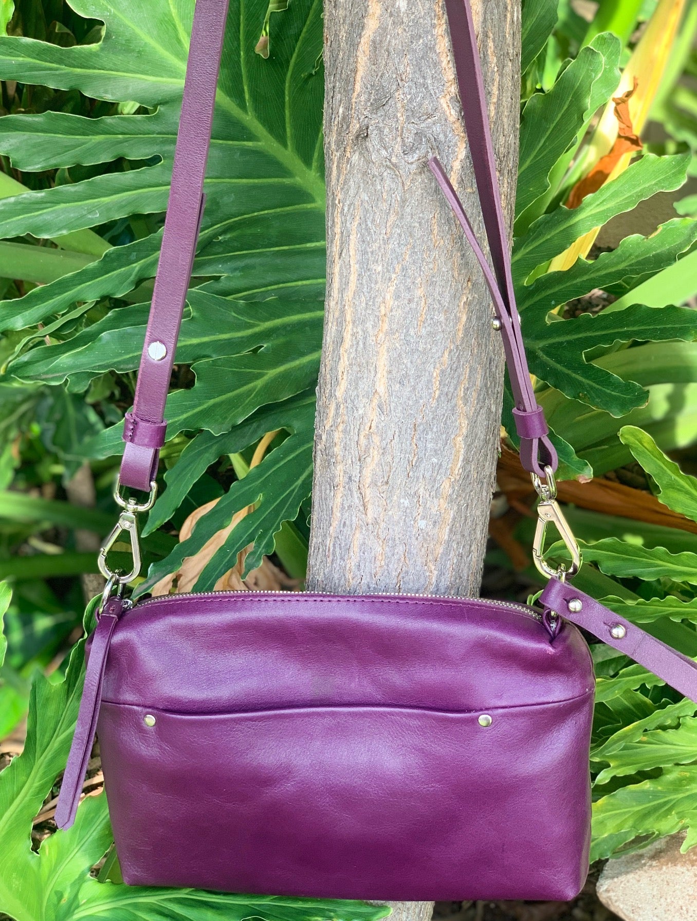 Plum purple shenn leather bag with a strap and a front pocket hang on a tree branck and green leaves in the background