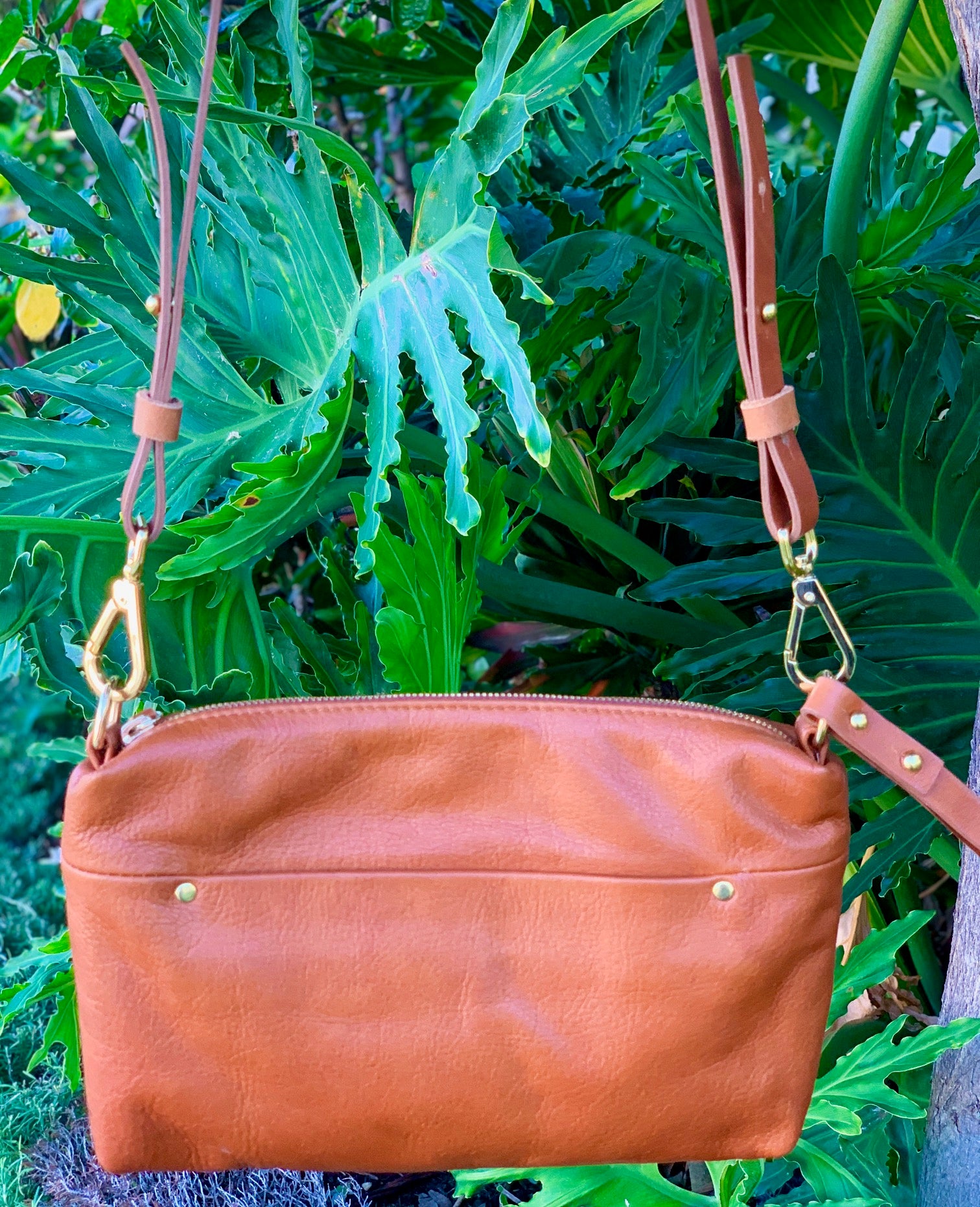 Whisky brown butter leather bag front view with a strap and a front pocket against a green leafy background