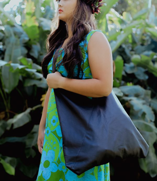 A woman wearing a green and blue floral dress holding a black lava leather bag as a crossbody bag against a greenery background.