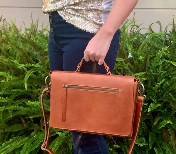 A woman holding a whisky brown leather handbag in front of green foliage.