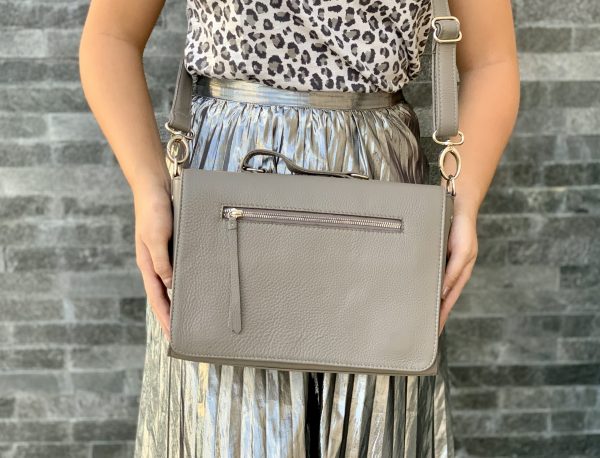 A woman holding a warm grey silky textured leather bag as a crossbody against a brick wall background