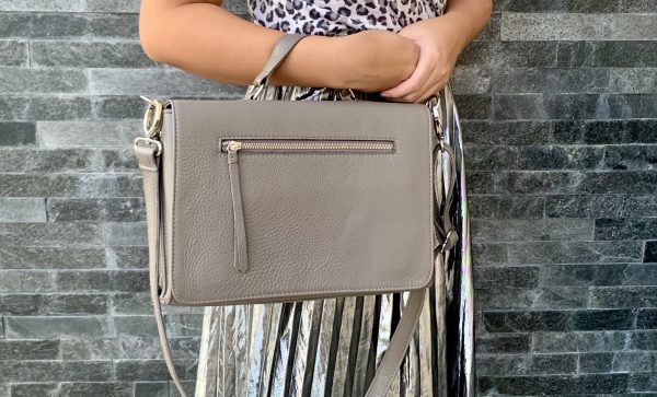 A woman holding a warm grey silky textured leather bag  in the crook of her arm against a brick wall.