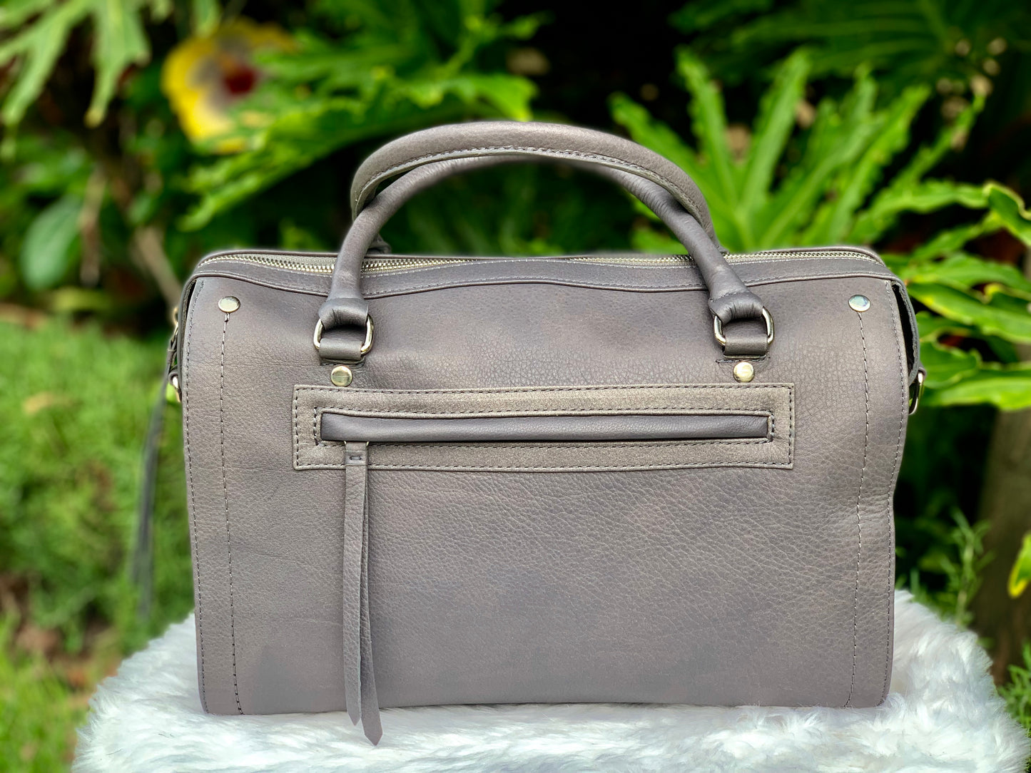 Titanium gray leather bag back view with an external zip pocket, on a white surface with green foliage in the background