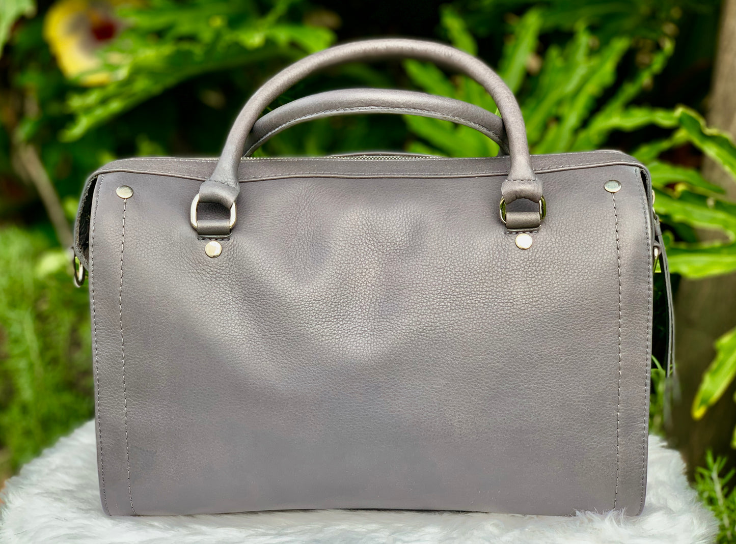 Titanium Gray leather bag front view on a white surface with green foliage in the background