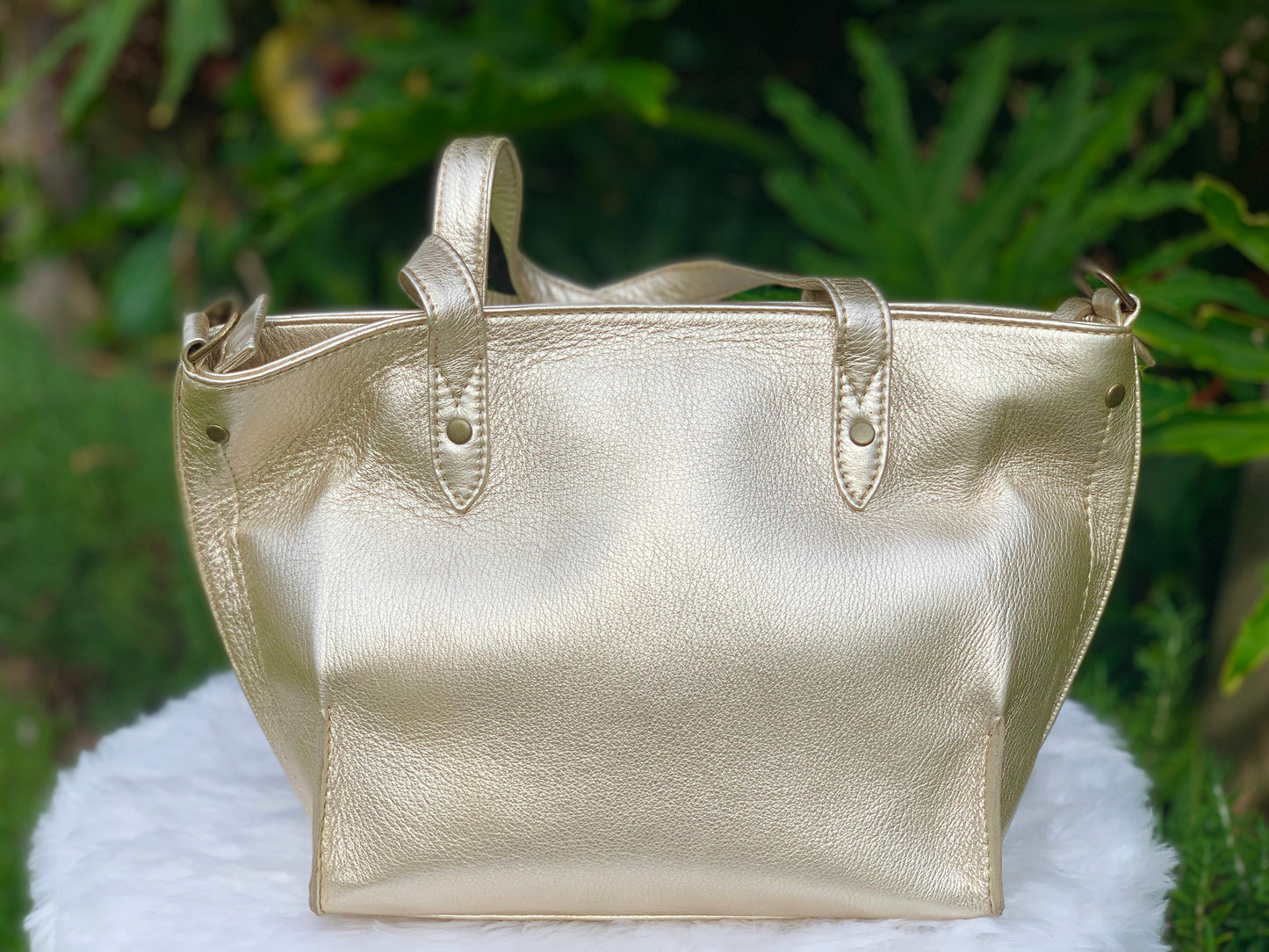 Pale gold metallic leather tote bag on a white surface with a green leafy background