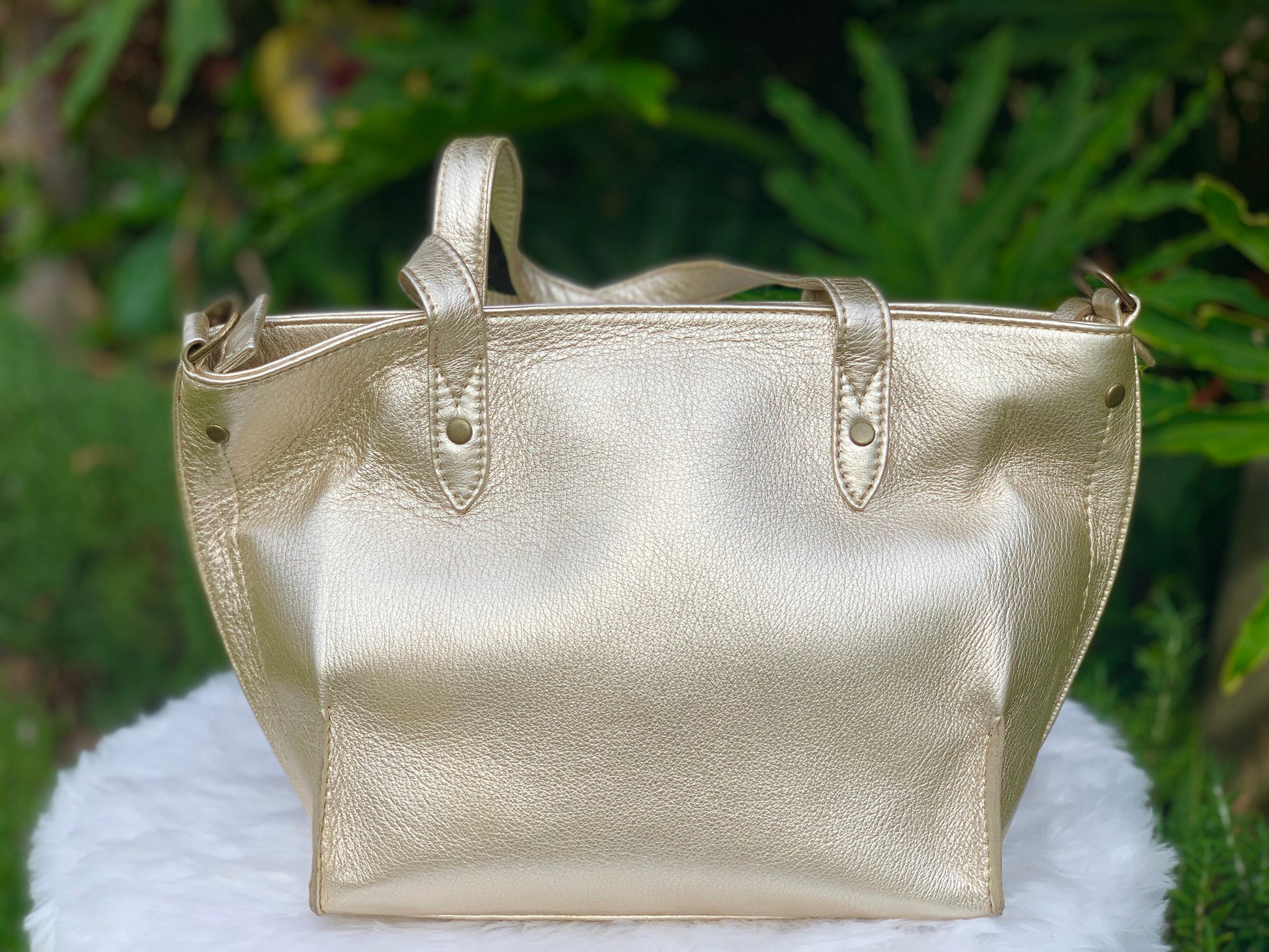 Pale gold metallic leather tote bag on a white surface with a green leafy background
