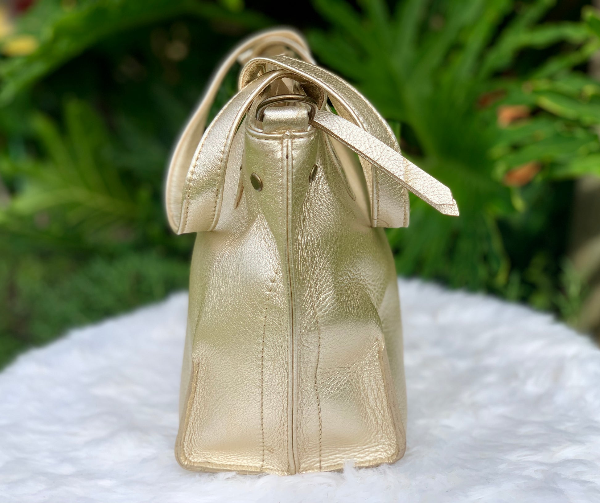 Pale gold metallic leather tote bag side view on a white surface with a green leafy background