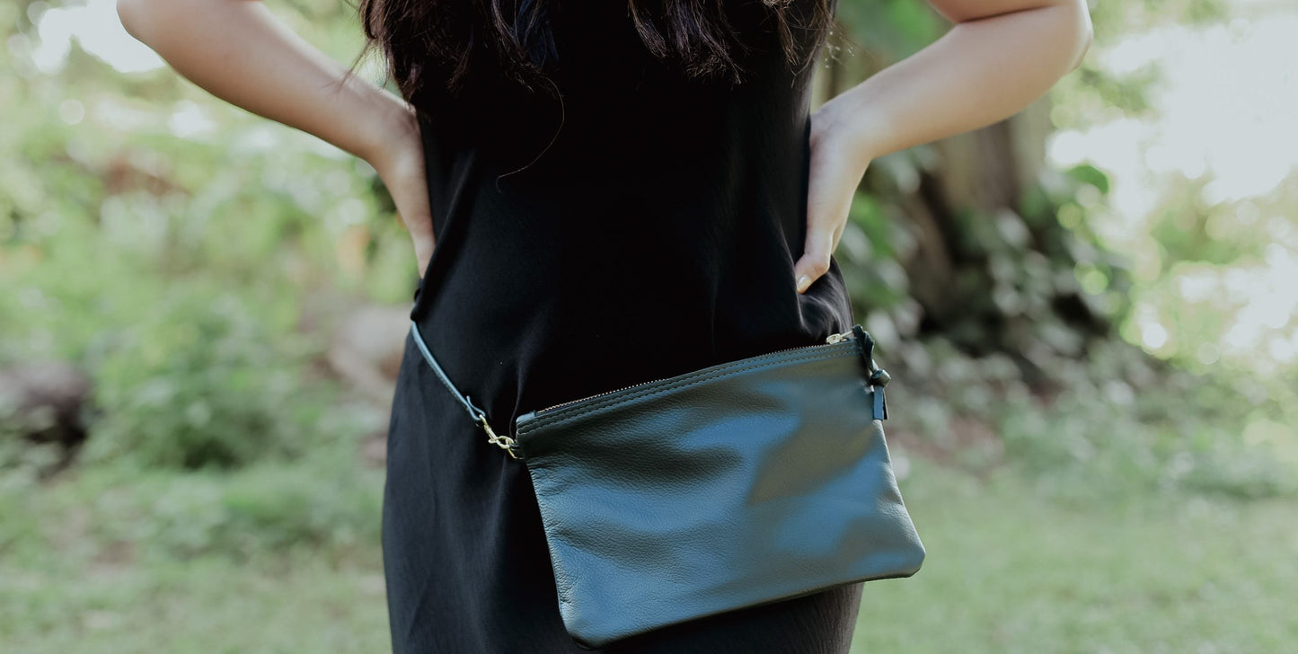 Close-up of a woman in a black dress wearing a sleek black leather pouch as a belt bag around her waist, standing in a lush green outdoor setting.