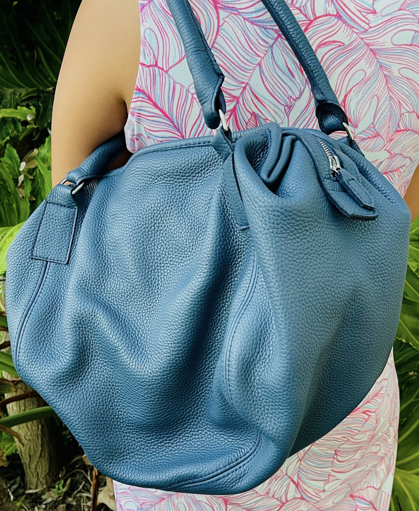 Denim Blue leather bag held by a person on the shoulder wearing a patterned dress against a natural greenery background