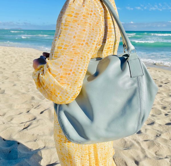 Woman holding a Light grey bag on the shoulder and wearing yellow patterned dress with a beach background.