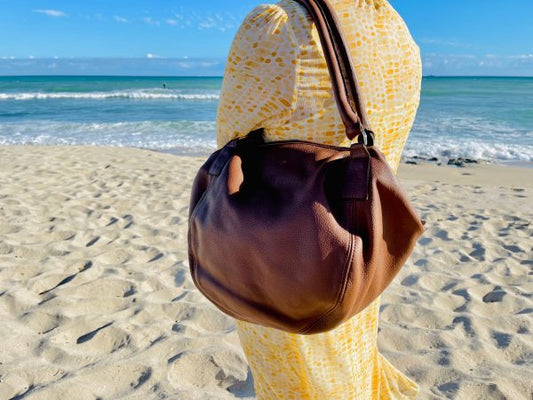 A woman wearing a yellow patterned dress, holding a large brown bag on their shoulder, standing on a sandy beach with the ocean in the background.
