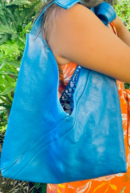 A woman holding on her shoulder a cobalt blue leather hobo bag with a tied strap against a greenery background. made in hawaii.