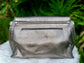 Pewter Metallic bag back view on a white surface with green leaves in the background