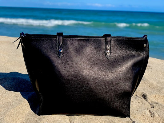 Onyx Black leather tote bag front view on a sandy beach with ocean in the background