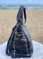 Sapphire leather satchel bag side view with gold studs and tassels on a fluffy white surface with a beach background