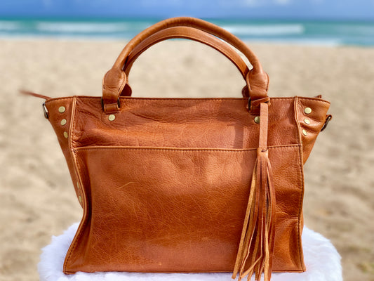 Whisky Brown leather satchel bag with tassels on a fluffy white surface with a beach background