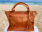 Whisky Brown leather satchel bag with tassels on a fluffy white surface with a beach background