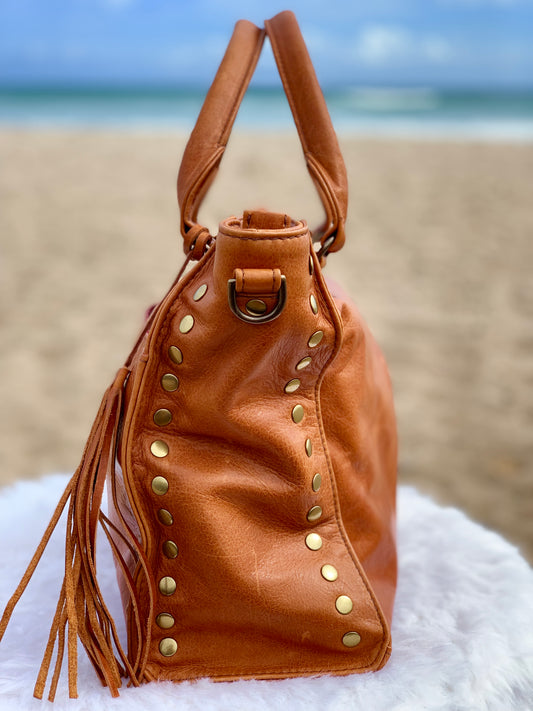 Whisky Brown leather satchel bag side view with gold studs and tassels on a fluffy white surface with a beach background