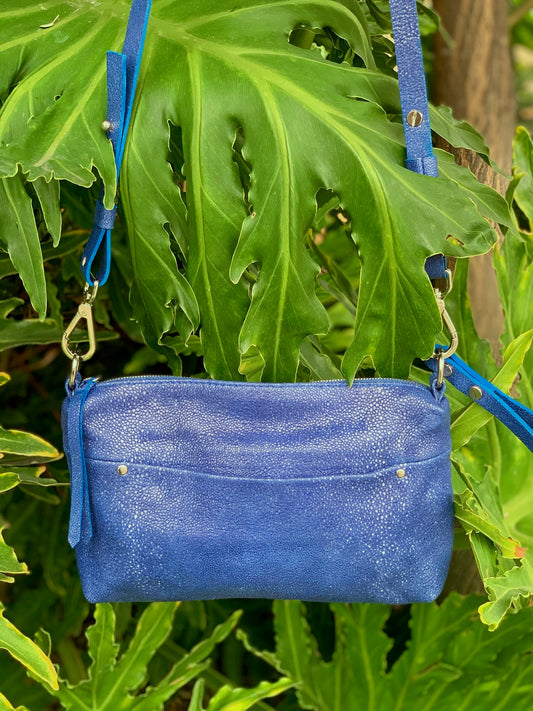 Ehukai Blue leather bag with a strap,  Front external slip pocket and Wrist strap against a green leafy background