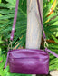 Plum purple shenn leather bag with a strap and a front pocket hang on a tree branck and green leaves in the background