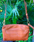 Whisky brown butter leather bag front view with a strap and a front pocket against a green leafy background