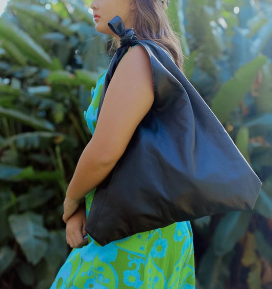 A woman wearing a green and blue floral dress holding a black lava leather bag  on her shoulder with a tied strap against a greenery background.