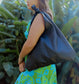 A woman wearing a green and blue floral dress holding a black lava leather bag  on her shoulder with a tied strap against a greenery background.