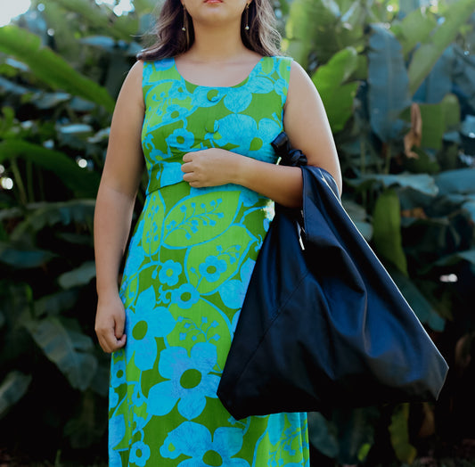 A woman wearing a green and blue floral dress holding a black lava leather bag in the crook of her arm against a greenery background.