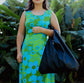 A woman wearing a green and blue floral dress holding a black lava leather bag in the crook of her arm against a greenery background.