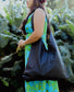 A woman wearing a green and blue floral dress holding a black lava leather bag on her shoulder with untied strap against a greenery backgroud.