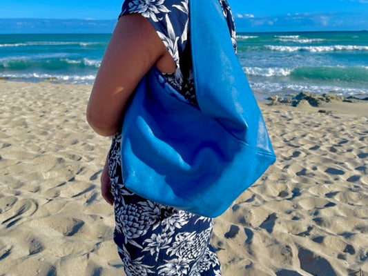 A woman holding on her shoulder a cobalt blue leather hobo bag with a tied strap on a sandy beach with ocean waves in the background. made in hawaii.