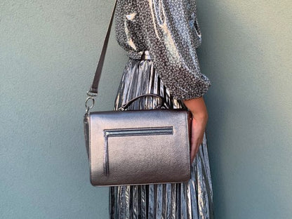 A woman holding a pewter metallic leather handbag on her shoulder against a plain background