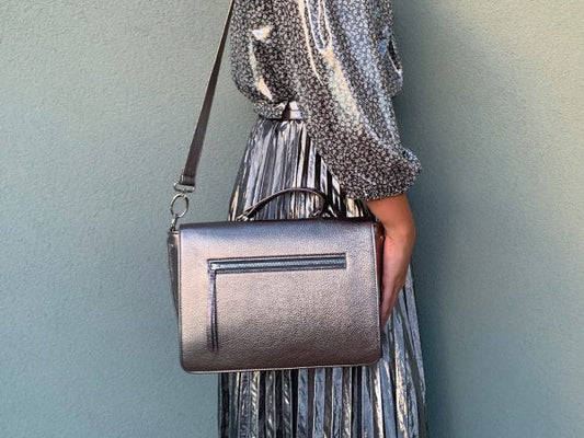 A woman holding a pewter metallic leather handbag on her shoulder against a plain background