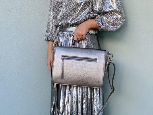 A woman wearing a silver sequin outfit holding a matching Pewter Metallic leather handbag against a light blue background