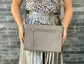 A woman holding a warm grey silky textured leather bag as a crossbody against a brick wall background