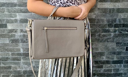 A woman holding a warm grey silky textured leather bag  in the crook of her arm against a brick wall.