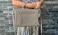 A woman holding a warm grey silky textured leather bag  in the crook of her arm against a brick wall.