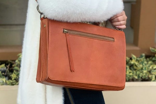 A whisky Brown leather bag held by a woman on her shoulder who's wearing a white fur coat.