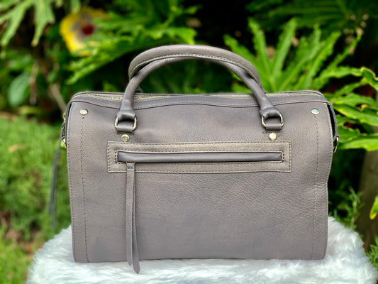 Titanium gray leather bag back view with an external zip pocket, on a white surface with green foliage in the background