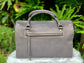 Titanium gray leather bag back view with an external zip pocket, on a white surface with green foliage in the background