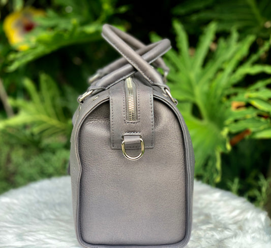 Titanium gray leather bag side view with a silver zipper on a white surface with green foliage in the background