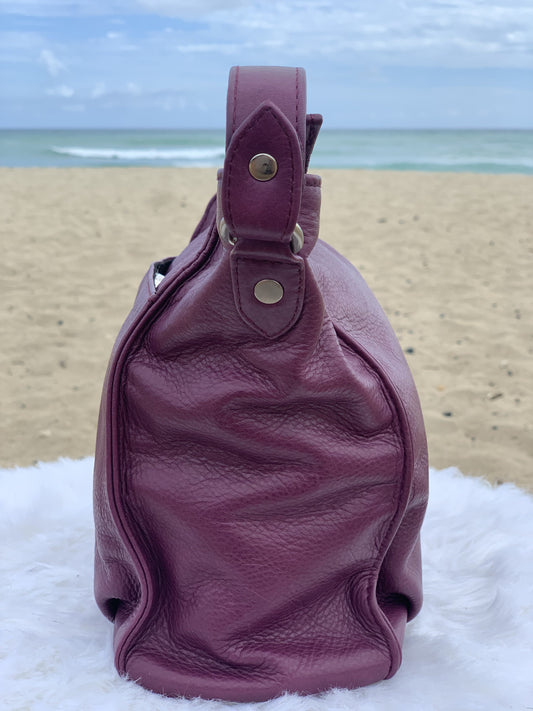 An aubergine hobo bag side view on a white surface with a beach background
