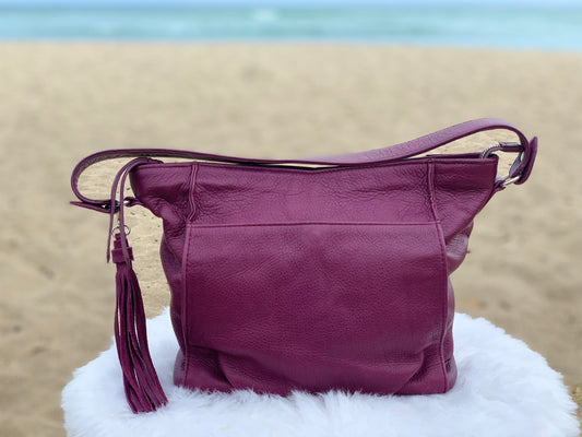 An aubergine hobo bag with a tassel on a white surface with a beach background