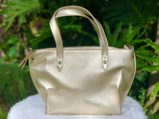 Pale gold metallic leather tote bag back view on a white surface with a green leafy background